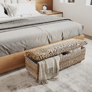 Könnte beinhalten: Eine rechteckige Aufbewahrungsbank mit einem Bezug in Leopardenmuster in Beige, Schwarz und Braun. Die Bank verfügt über einen teilweise geöffneten Klappdeckel, der einen hohlen Innenraum freigibt, und eine beige Decke mit Fransen hängt vorne über.
