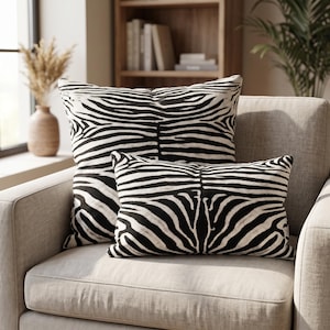 Peut inclure: Deux coussins décoratifs arborant un motif de rayures zébrées noires et blanches. L'un est carré et l'autre est rectangulaire. Les deux coussins présentent un design symétrique cohérent avec des rayures sombres sur un fond blanc cassé.