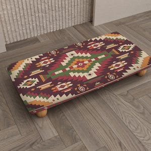 May include: A white ceramic mug with a black handle and the text "But First, Coffee" in a bold, black font. The mug is set against a blurred background, suggesting a cozy setting.