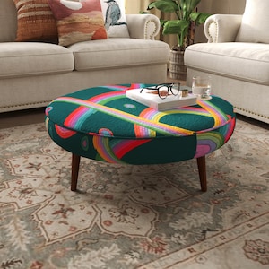 May include: A round ottoman featuring a dark teal fabric base with a vibrant, multi-colored pattern of concentric rainbow-hued arches and circular shapes. The piece is supported by four tapered wooden legs and holds a white book and glasses.