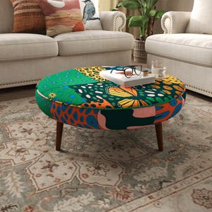 May include: A round ottoman featuring a vibrant patchwork pattern of green, orange, and dark teal with butterfly, leopard print, and floral motifs. The piece stands on four tapered wooden legs and holds a white book with black glasses on top.