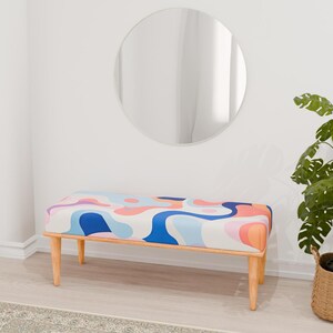 May include: A wooden bench with a colorful abstract pattern on the seat cushion. The bench has a light wood finish and is sitting on a light gray rug in front of a white wall. A round mirror and a potted plant are visible in the background.