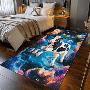 Op de afbeelding: Een rechthoekig tapijt met een achtergrond van een sterrenhemel. Een zwart-witte koe zweeft in de ruimte met planeten en sterren eromheen. Het tapijt heeft een rechthoekige vorm en is in blauw, paars en roze tinten.