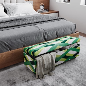 May include: A modern bedroom scene featuring a bed with a gray duvet and pillows. A geometric patterned storage bench in green, white, and black sits at the foot of the bed, with a blanket draped over the edge. A gray rug covers the floor.