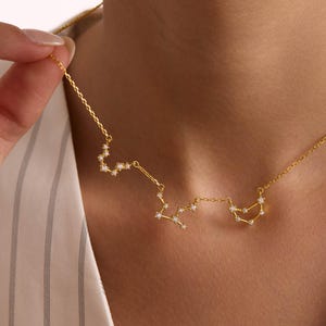 May include: A gold-colored necklace featuring three zodiac constellations: Scorpio, Sagittarius, and Capricorn. The necklace has a delicate chain with small, sparkling accents representing stars. The constellations are connected by fine gold chains.