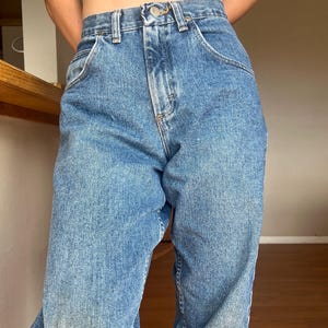 May include: Light blue denim jeans with a high waist and classic five-pocket design. The jeans have a straight leg cut and a button and zipper closure. The denim has a faded wash and some subtle wear marks, giving them a vintage look.