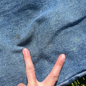 May include: Close-up of light blue denim fabric with a few dark spots and a hand making a peace sign. The denim has a textured appearance and is slightly wrinkled. The background is green grass.