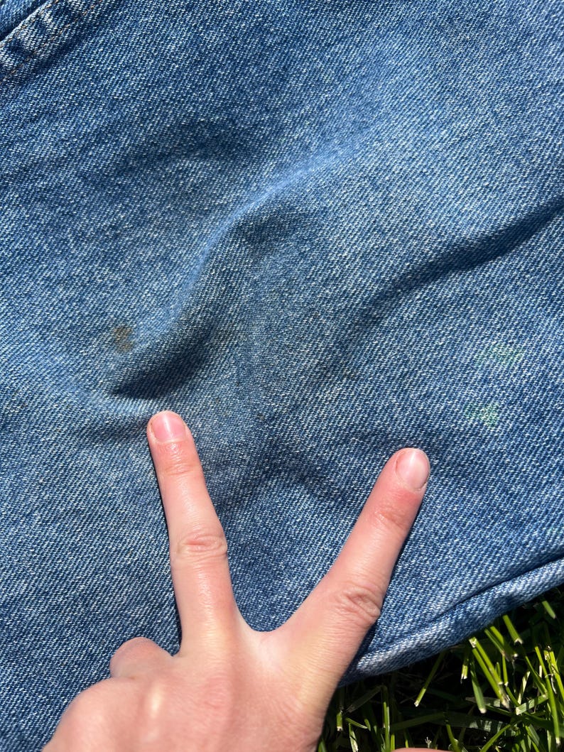May include: Close-up of light blue denim fabric with a few dark spots and a hand making a peace sign. The denim has a textured appearance and is slightly wrinkled. The background is green grass.