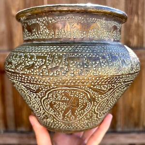 May include: A round brass pot featuring intricate hand-engraved patterns. The surface displays horizontal bands of stylized animals and geometric motifs, with a central cartouche depicting a figure holding a staff surrounded by birds.
