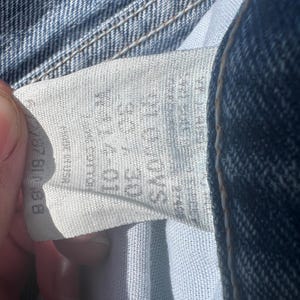 May include: Close-up of a light blue denim jeans label. The label displays size information, including a waist size of 30 inches. The jeans are made of cotton. The label is partially visible, showing the brand and care instructions.