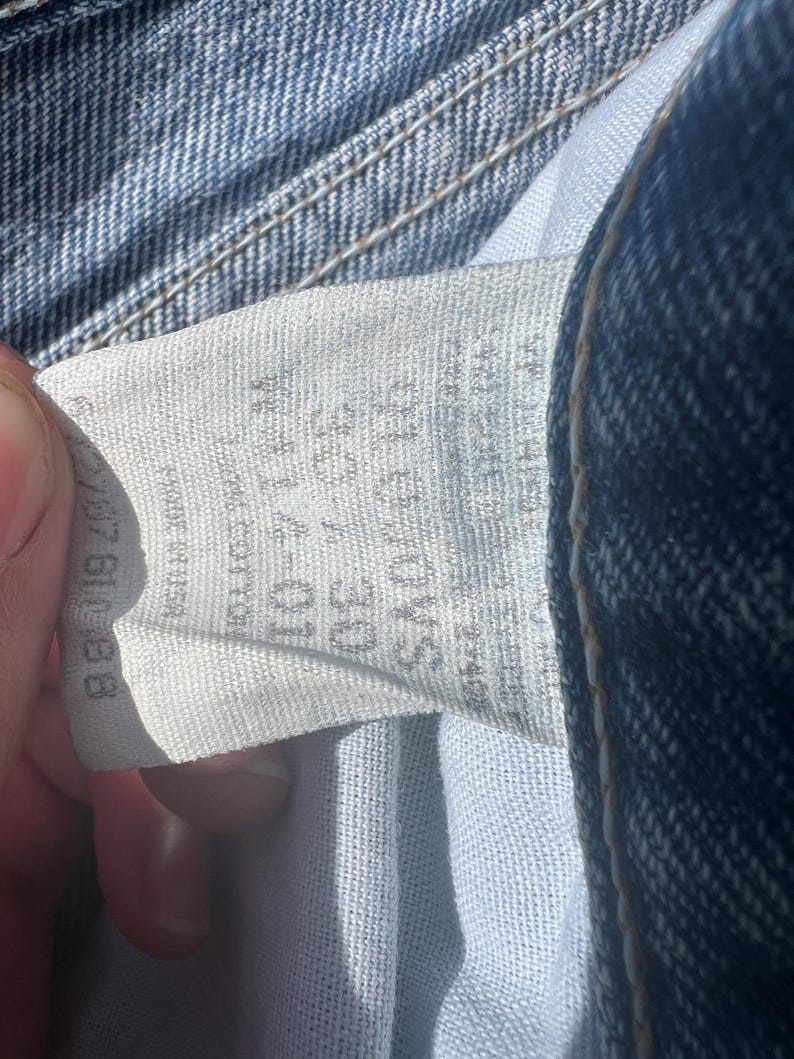 May include: Close-up of a light blue denim jeans label. The label displays size information, including a waist size of 30 inches. The jeans are made of cotton. The label is partially visible, showing the brand and care instructions.