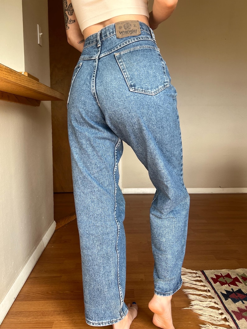 May include: Light blue denim Wrangler jeans. The jeans have a high waist and a straight leg fit. The Wrangler logo is visible on the back waistband. The jeans are worn with a light-colored top.