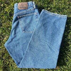May include: Blue denim Wrangler jeans with a classic five-pocket design. The jeans feature a brown leather patch with the Wrangler logo on the back waistband. The jeans are laid out on a bed of green grass.