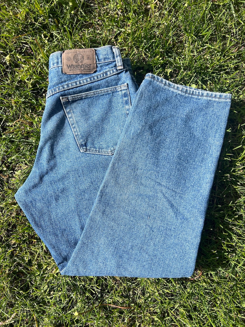 May include: Blue denim Wrangler jeans with a classic five-pocket design. The jeans feature a brown leather patch with the Wrangler logo on the back waistband. The jeans are laid out on a bed of green grass.