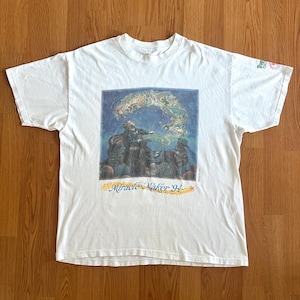 May include: A white short-sleeve t-shirt featuring a central graphic of a person gesturing toward a swirling celestial scene with animals. The text "Miracle Maker '94" is printed in yellow below the image, and a small logo is on the left sleeve.