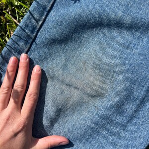 May include: Close-up of light blue denim jeans with a faded area. The jeans are lying on green grass. A hand is partially visible, resting on the denim. The jeans show signs of wear and tear.