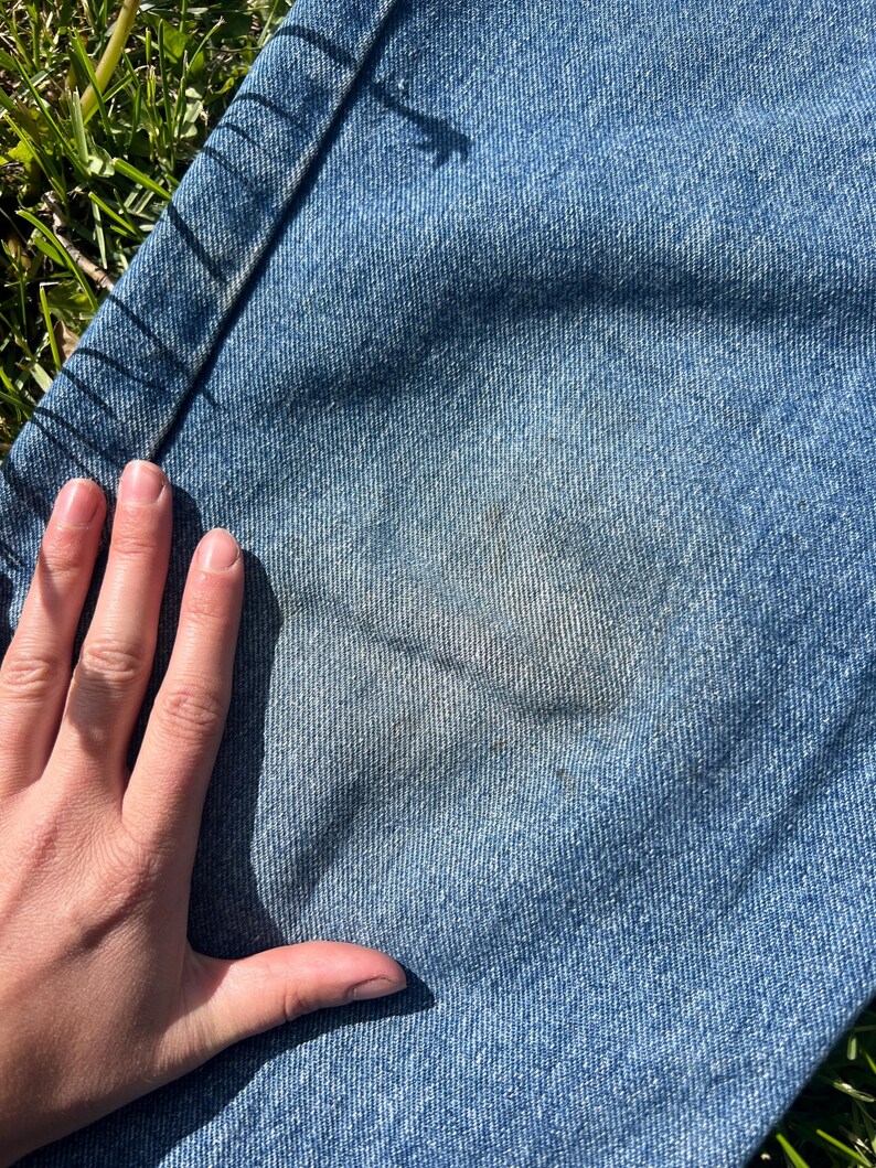 May include: Close-up of light blue denim jeans with a faded area. The jeans are lying on green grass. A hand is partially visible, resting on the denim. The jeans show signs of wear and tear.