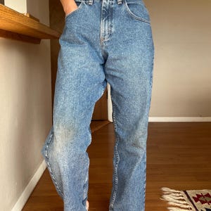 May include: Light blue denim jeans with a classic five-pocket design. The jeans have a straight leg cut and a button and zipper closure. The denim has a faded wash, giving them a vintage look. The jeans are worn at the knees.