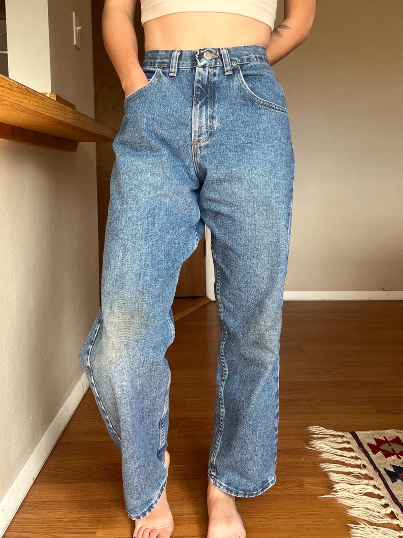 May include: Light blue denim jeans with a classic five-pocket design. The jeans have a straight leg cut and a button and zipper closure. The denim has a faded wash, giving them a vintage look. The jeans are worn at the knees.
