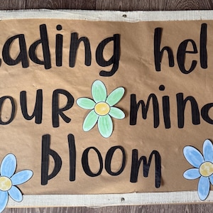 May include: A brown banner with the words "reading helps your mind bloom" in black lettering. The banner is decorated with colorful paper flowers in shades of blue, purple, and green. Green leaves are also visible.