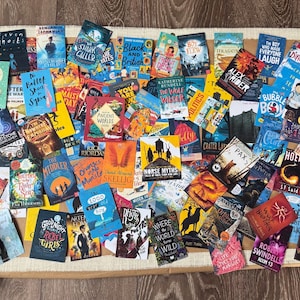 May include: A selection of book covers, such as "Ella", "Survivors", and "PAX", in various colours, arranged on a neutral surface. The books vary in size and design, creating a visually interesting arrangement.