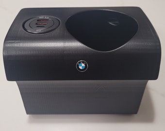 May include: Black car console with a cup holder and a charging port. The charging port has the text "QC 3.0" and "PD". A BMW logo is on the front.