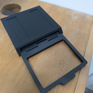 May include: A black plastic case with a hinged lid that opens to reveal a square frame. The case is designed to hold and display a photograph or other artwork.