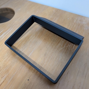 Może przedstawiać: Czarna, kwadratowa metalowa rama z płaskim wierzchem, spoczywająca na jasnej drewnianej powierzchni. Rama ma prosty, nowoczesny design z czystymi liniami i matowym wykończeniem. Prawdopodobnie jest to przedmiot dekoracyjny lub funkcjonalny.