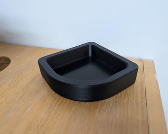 May include: A black, corner-shaped tray with a smooth interior and a slightly textured exterior. The tray is made of a solid material and sits on a light-colored wooden surface. The design is simple and modern.