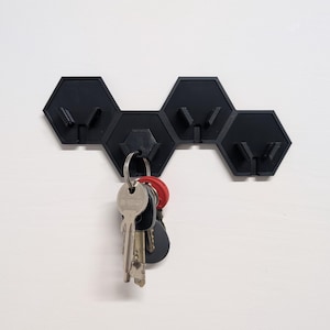 May include: Black hexagonal key holder with multiple hooks, mounted on a white wall. A set of keys is hanging from one of the hooks.