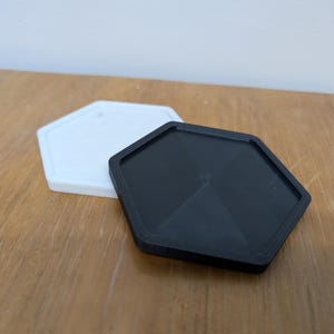 May include: Two hexagonal coasters, one white and one black. The black coaster has a raised, star-shaped design in the centre.