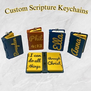 May include: Five custom scripture keychains with different names and bible verses. The keychains are made of a dark blue material with gold lettering. The keychains are in the shape of a small book. The keychains are arranged in a semi-circle on a white surface.