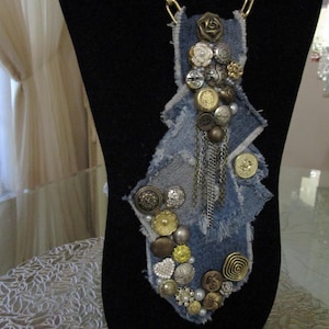 May include: A necklace featuring a blue denim fabric base with frayed edges, adorned with an assortment of metallic and decorative buttons in various shapes and sizes, including a dangling silver chain element and a gold-toned metal chain.