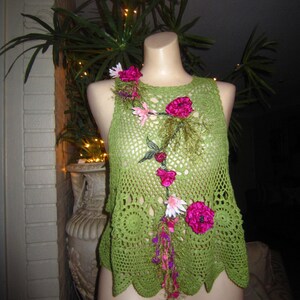 May include: Green crochet top with a floral design featuring pink and white flowers. The top has a scalloped hem and a loose fit.