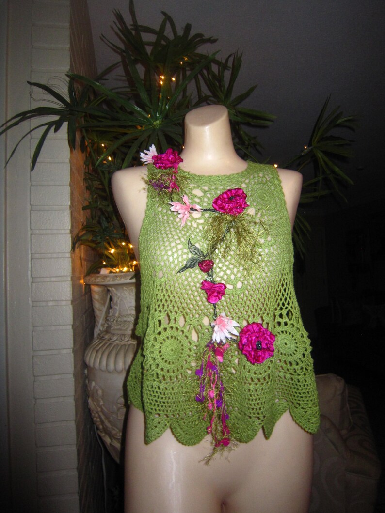 May include: Green crochet top with a floral design featuring pink and white flowers. The top has a scalloped hem and a loose fit.