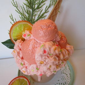 May include: A pink ice cream sundae with whipped cream, sprinkles, and orange slices. The sundae is in a waffle cone and is sitting on a white doily on a green plate.