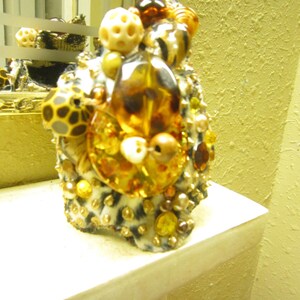 May include: A decorative object with a leopard print base, adorned with various beads and gems in shades of amber, gold, and cream. The object is placed on a white shelf, with a textured yellow wall in the background. The design incorporates a variety of shapes and textures.