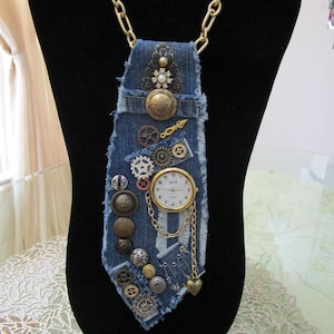 May include: A denim necktie accessory featuring a frayed edge design, embellished with various brass-toned metal gears, assorted circular buttons, safety pins, and a central analog watch face with a gold-toned chain and a small heart charm.