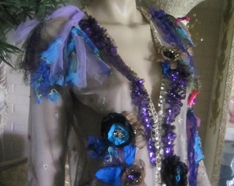 May include: A sheer, light brown top with a plunging neckline and a layered design. The top is adorned with blue, purple, and pink fabric flowers and sequins.