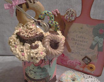 May include: A pink and white ceramic mug decorated with a cupcake, donut, and other sweets. The mug is filled with whipped cream, chocolate sprinkles, and a silver spoon. The mug is sitting on a pink and white cutting board with the text "Cafés, Chocolat, Pâtes et Sirops".