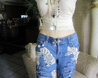 May include: A pair of blue denim shorts with white crochet detailing. The shorts are ripped and distressed. The model is wearing a white lace tank top with a layered pearl and silver necklace and a black flower brooch.