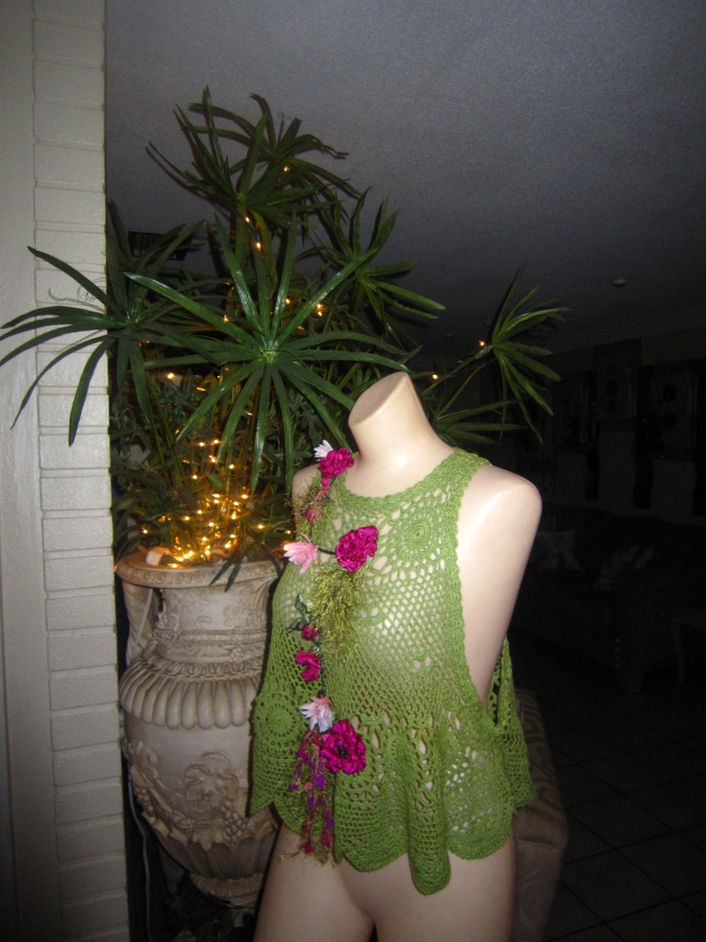 May include: Green crochet top with pink flower embellishments. The top has a loose, flowy fit and a scalloped hem.