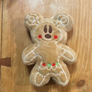 K&ouml;nnte beinhalten: Ein Pl&uuml;sch-Lebkuchenmann-Ornament mit Mickey Mouse-Ohren. Das hellbraune Ornament hat wei&szlig;e N&auml;hte, ein braunes Gesicht, rote Wangen und eine Halskette aus roten und gr&uuml;nen Punkten. Das Ornament befindet sich auf einer Holzoberfl&auml;che.