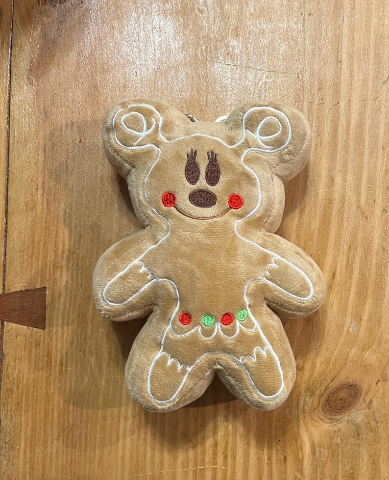 K&ouml;nnte beinhalten: Ein Pl&uuml;sch-Lebkuchenmann-Ornament mit Mickey Mouse-Ohren. Das hellbraune Ornament hat wei&szlig;e N&auml;hte, ein braunes Gesicht, rote Wangen und eine Halskette aus roten und gr&uuml;nen Punkten. Das Ornament befindet sich auf einer Holzoberfl&auml;che.