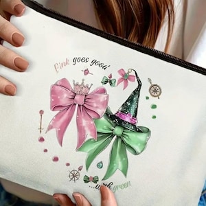 May include: A white canvas cosmetic bag with a black zipper, featuring a whimsical design. The design includes a pink bow with a crown, a green witch hat, and the text "Pink goes good...with green."