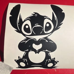 May include: Black vinyl decal of the character Stitch from Lilo & Stitch. The cartoon alien is depicted with large eyes, a heart-shaped nose, and a heart formed by his hands. The decal is on a white background.