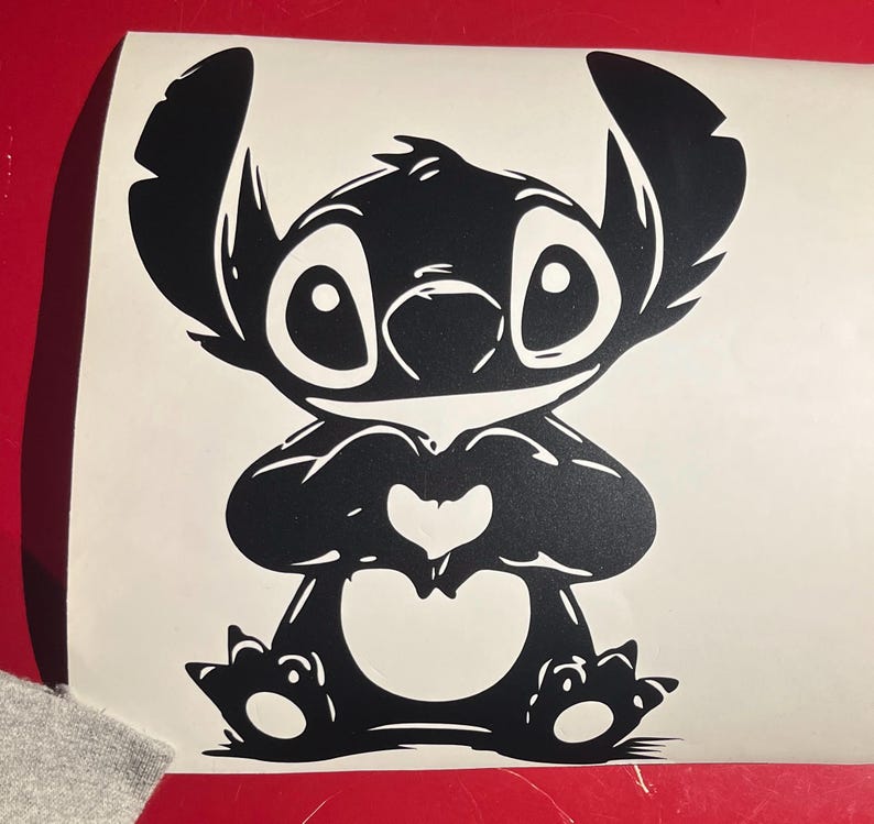 May include: Black vinyl decal of the character Stitch from Lilo & Stitch. The cartoon alien is depicted with large eyes, a heart-shaped nose, and a heart formed by his hands. The decal is on a white background.