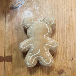 K&ouml;nnte beinhalten: Ein Pl&uuml;schspielzeug in Form eines Lebkuchenmanns in hellbrauner Farbe mit wei&szlig;en N&auml;hten, die die Form umrei&szlig;en. Das Spielzeug liegt auf einer Holzoberfl&auml;che.