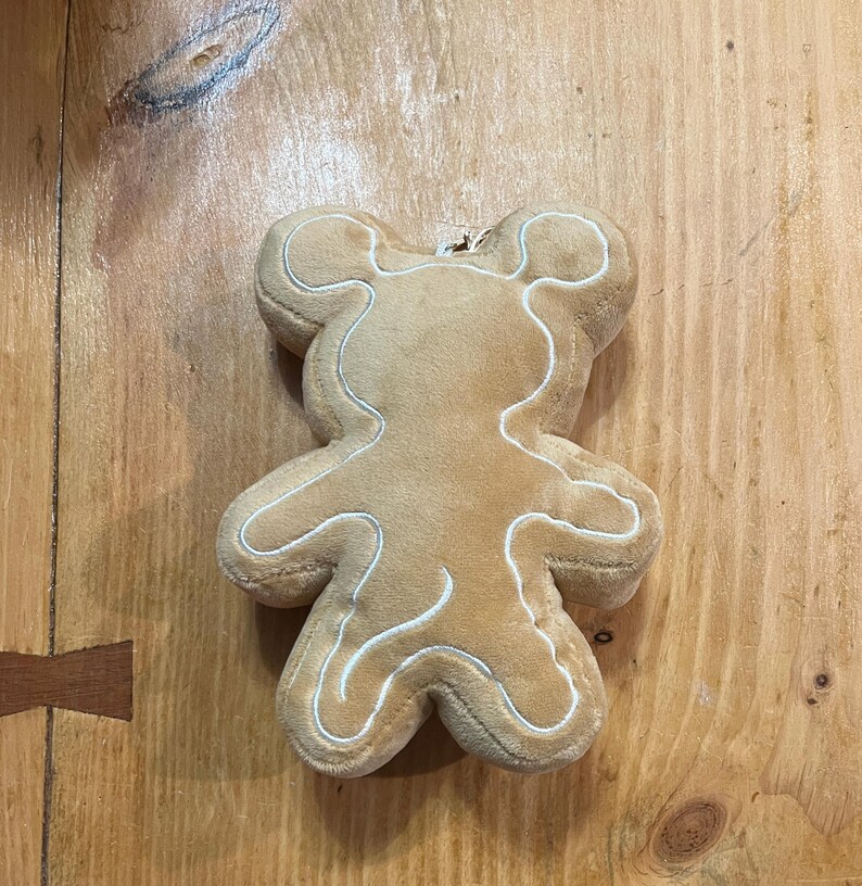 K&ouml;nnte beinhalten: Ein Pl&uuml;schspielzeug in Form eines Lebkuchenmanns in hellbrauner Farbe mit wei&szlig;en N&auml;hten, die die Form umrei&szlig;en. Das Spielzeug liegt auf einer Holzoberfl&auml;che.
