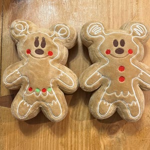 K&ouml;nnte beinhalten: Zwei Pl&uuml;sch-Lebkuchenm&auml;nner in Form von Spielzeugen mit Mickey Mouse-Ohren. Die Spielzeuge sind hellbraun mit wei&szlig;en Verzierungen, roten und gr&uuml;nen Akzenten und braunen Gesichtsz&uuml;gen. Die Spielzeuge liegen auf einer Holzoberfl&auml;che.